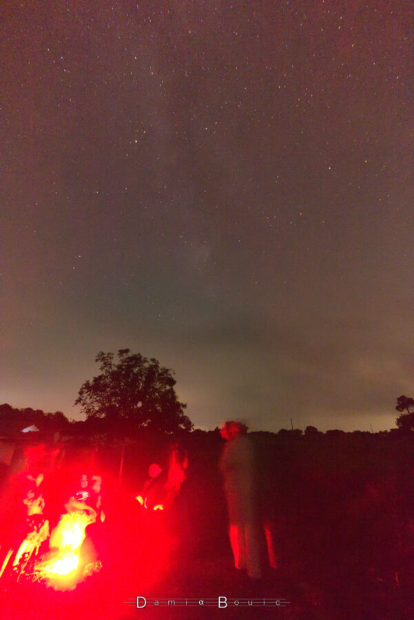 Scène nocturne façon feu de camps. Des gens sont regroupés autour mais on ne peut définir leur contours, floutés par l'exposition longue de la photo qui doit révéler le ciel étoilé au dessus de la scène ainsi que la Voie Lactée, que l'on parvient à identifier dans cette photo bruitée car ayant été prise avec les paramètres ISO les plus élevés