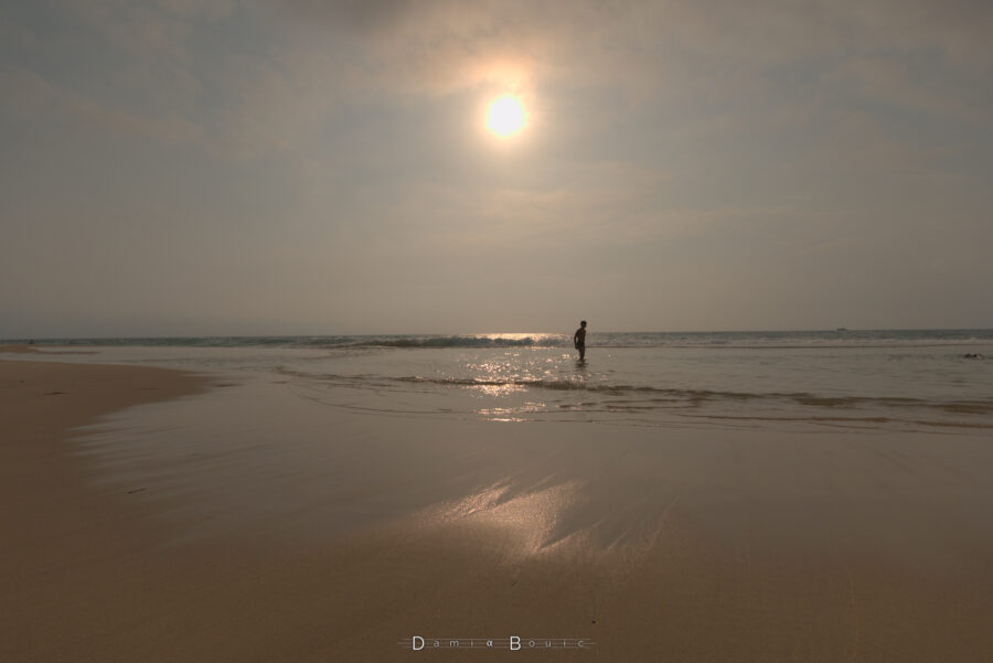 Soleil au dessus de l'océan qui reflète sa lumière sur l'eau et le sable lissé, doux, presque comme du velours. Une silhouette de baigneur se profile entre deux vagues sous le Soleil