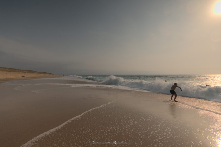 Cliché pris dans la perspective de la plage, offrant une vue sur une vague qui vient de s'abattre tout le long, se changeant en éclats de mousses et d'écumes. Un surfeur essaie de glisser sur la fine pellicule d'eau résiduelle sur le sable lissé en permanence par le resac de l'océan