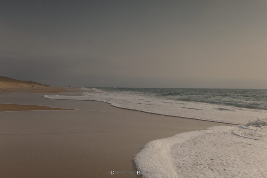 Photo prise au raz de l'écume qui finit sa course sur le sable lisse. La plage au dessus s'étire jusqu'à l'horizon sous un ciel typique d'une fin d'après-midi d'été, au couleurs chaudes.