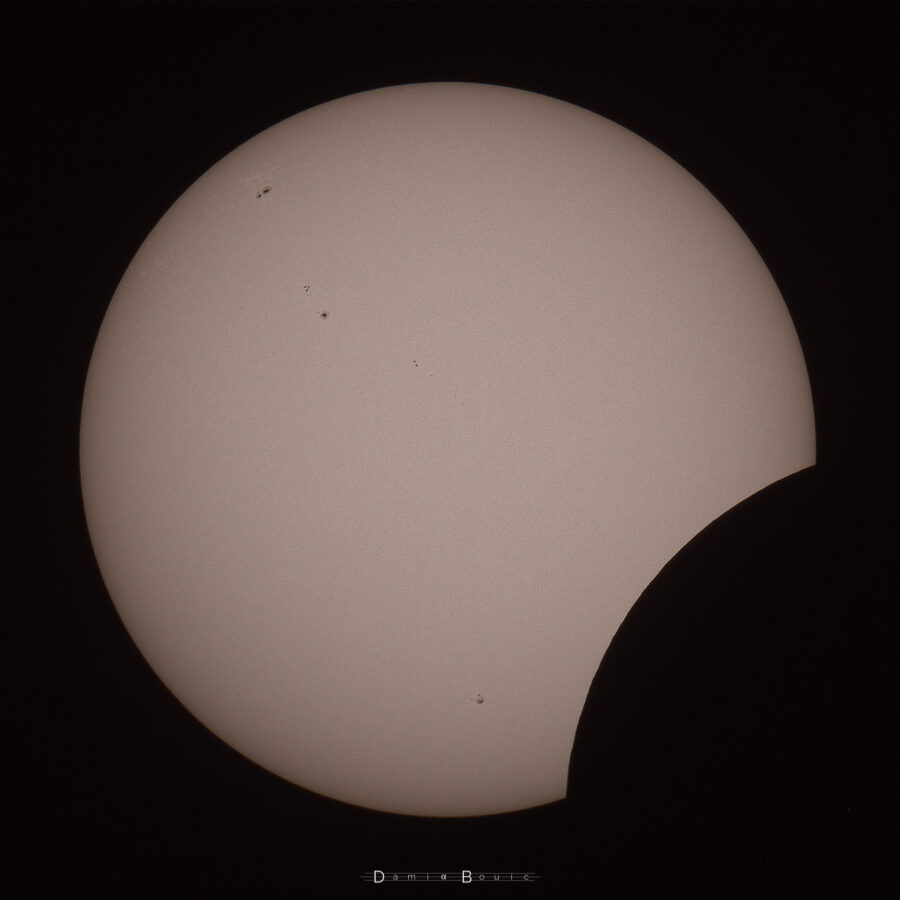 Le disque solaire, présentant de nombreuses tâches, notamment en haut à gauche et vers le milieu, qui commence à être recouvert par le disque lunaire, qui l'échancre en bas à droite.
